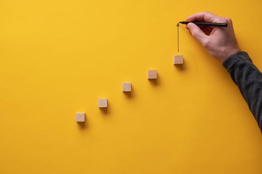 Male hand drawing an upward pointing arrow on top of a growing graph made of wooden blocks. Over yellow background.