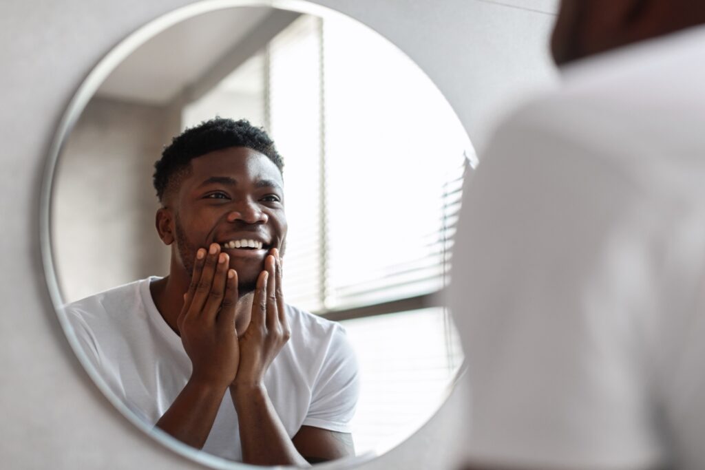 Happy African American Man Touching Unshaven Chin And Smiling To His Reflection In Round Mirror Standing In Bathroom At Home. Male Facial Skincare And Shaving Routine